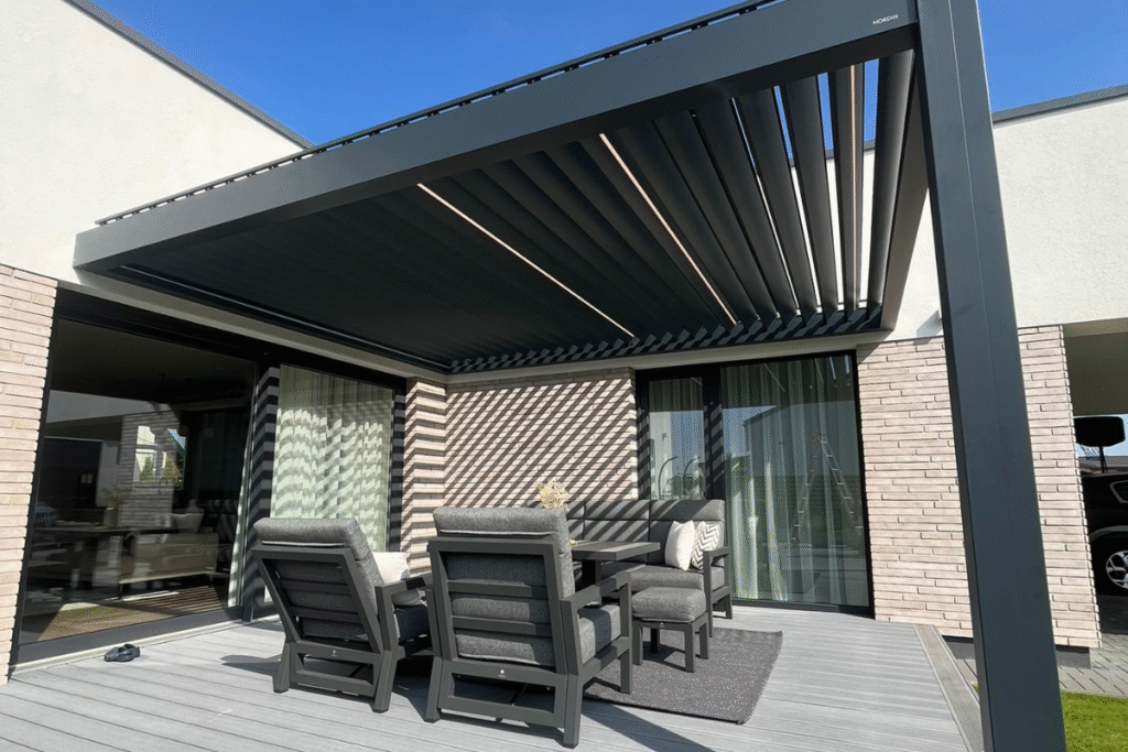 C7 Elite pergola su pakreipiamomis lamelėmis ir lauko baldais, įrengta prie šviesaus, modernios architektūros namo.