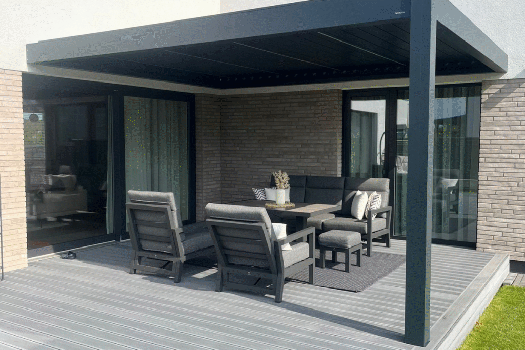 Ta pati pergola iš kito kampo – atvira, stilingai integruota į terasą su minkštais baldais ir stumdomais stiklais.